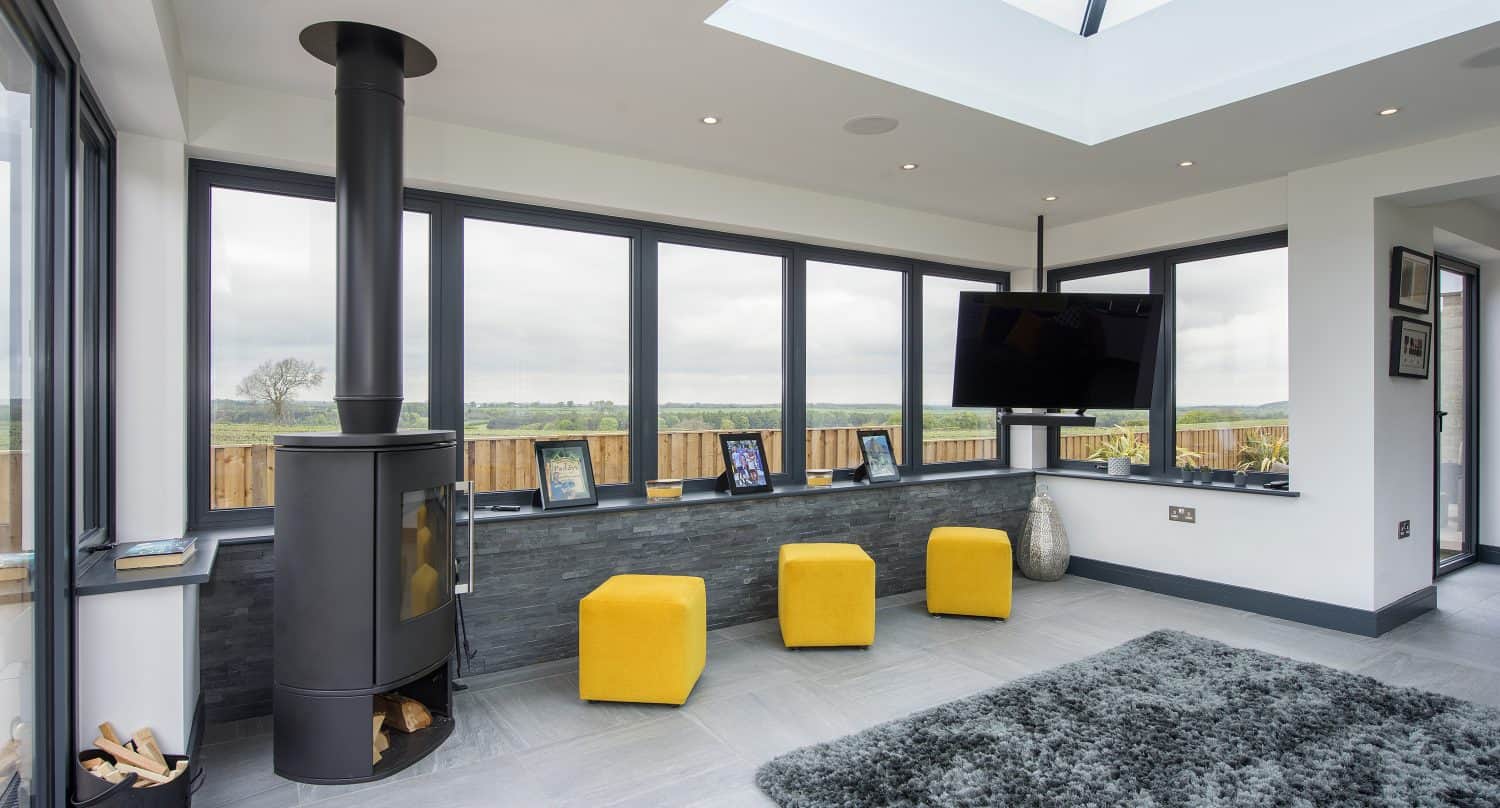 A modern sunroom with floor-to-ceiling windows and bespoke glass solutions, a black wood-burning stove, three yellow ottomans, a gray rug, wall-mounted TV, and framed photos on a low ledge, overlooking a green landscape.