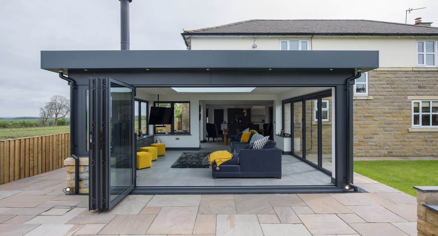 Modern home extension with large glass sliding doors and sleek glass roofing, opening to a living area with yellow and dark blue sofas, a rug, and a view into the dining space. Stone patio in the foreground and grassy yard beside.