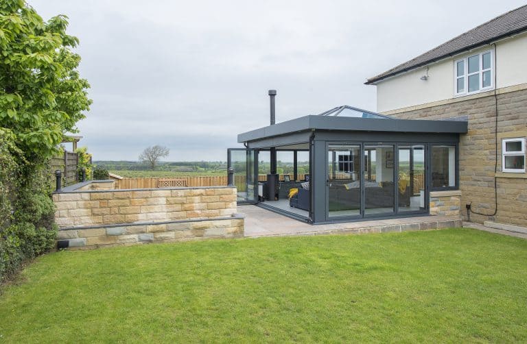 A modern glass conservatory with a black frame and bifold doors is attached to a stone and white house, overlooking a green lawn and countryside. The patio area features outdoor seating and a low stone wall.