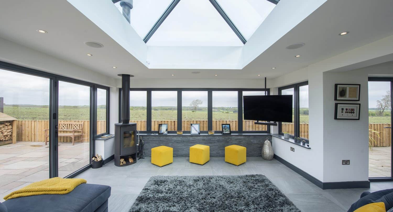 Modern living room with large glass windows, a skylight ceiling, and countryside views. The space features yellow stools, a gray rug, a sofa, TV, framed photos on a shelf, and sliding doors leading to the visible outdoor patio.