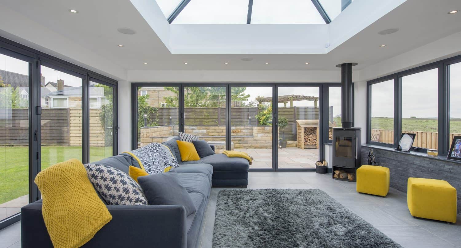 Modern sunroom with large glass roofing, grey sofas accented with yellow throws and cushions, grey rug, yellow stools, and floor-to-ceiling bifold doors opening to a patio and garden.