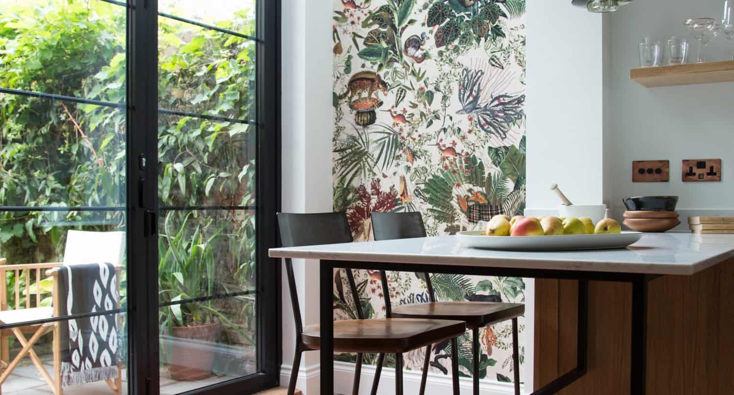 Modern kitchen with terracotta floor tiles, a marble island with black stools, glass pendant lights, and large bifold doors opening to a garden patio; one wall features vibrant botanical wallpaper.