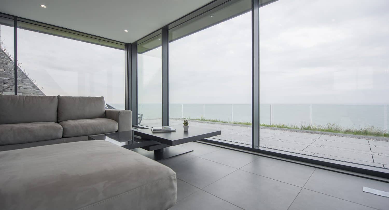 Modern living room with floor-to-ceiling glass windows and sliding doors offering a scenic view of the ocean. Minimalist decor features a light gray sofa, low black table, tiled floor, and an outdoor patio visible beyond the windows.