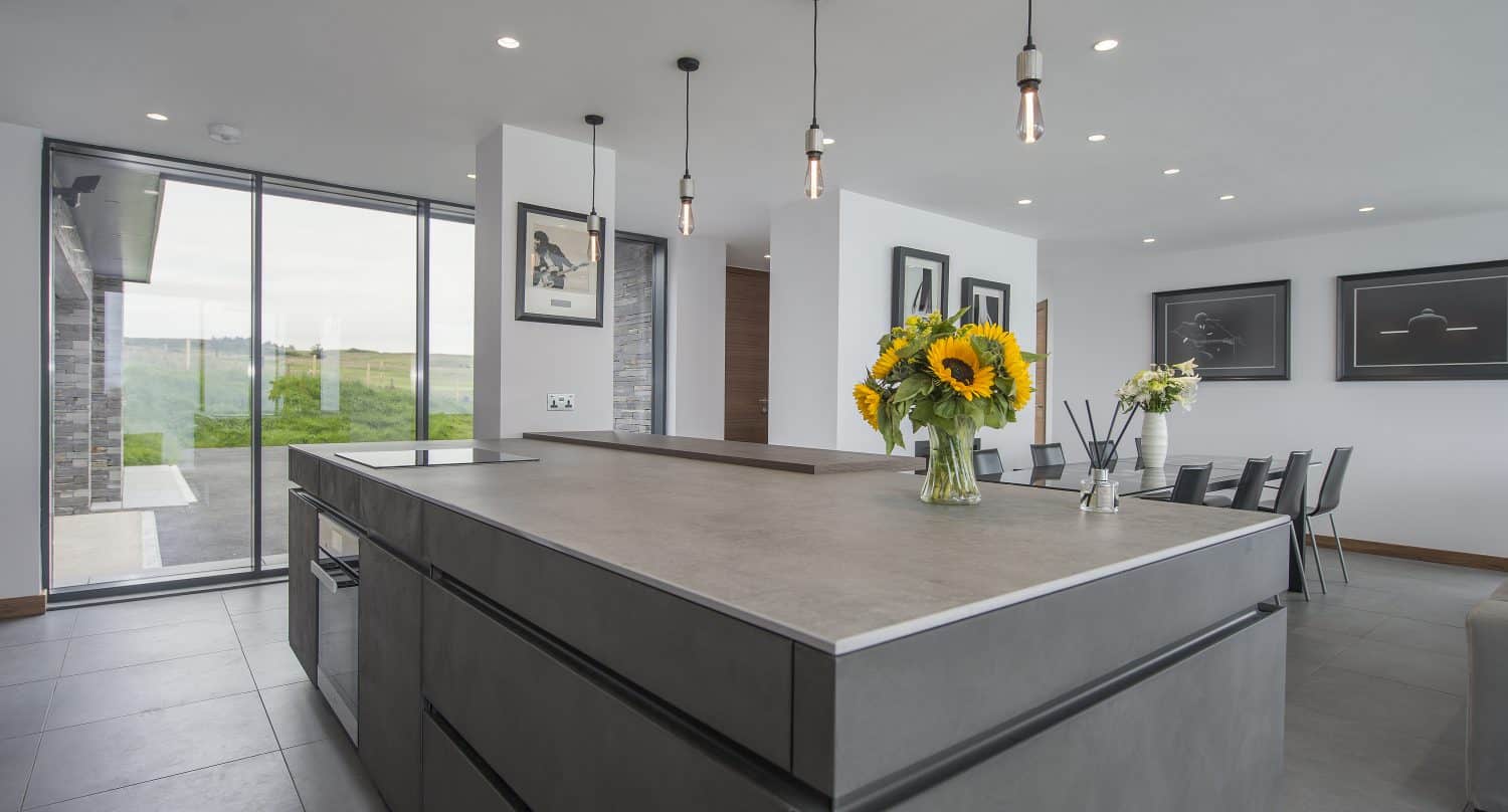 Modern kitchen with a large gray island, sunflowers in a vase, pendant lights above, and a dining table in the background. Floor-to-ceiling sliding doors let in natural light and offer views of greenery outside.