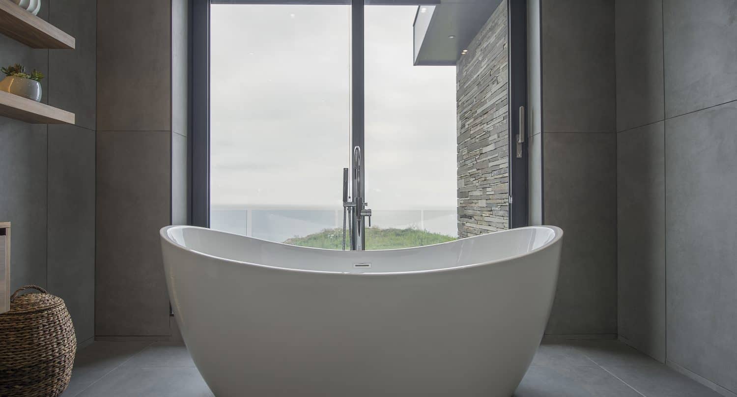 Modern bathroom with a white freestanding bathtub positioned in front of large sliding doors overlooking an outdoor view; the space has gray tiles, shelves with decor, and a woven basket. Bespoke glass solutions enhance the room’s contemporary elegance.