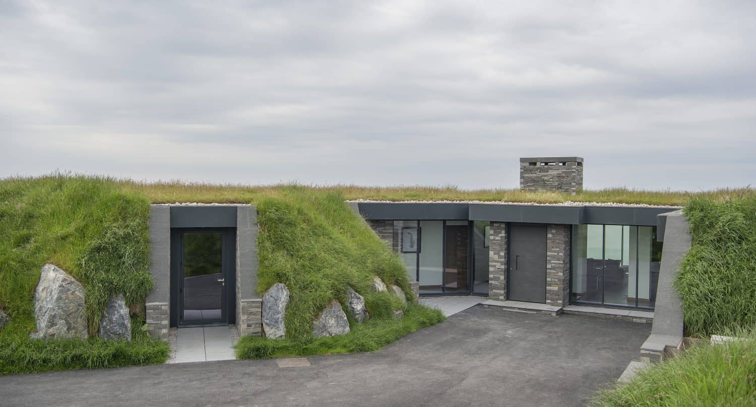 Modern house built into a grassy hillside, with large windows, sliding doors, and stone accents, partially covered by earth and surrounded by green grass under a cloudy sky.