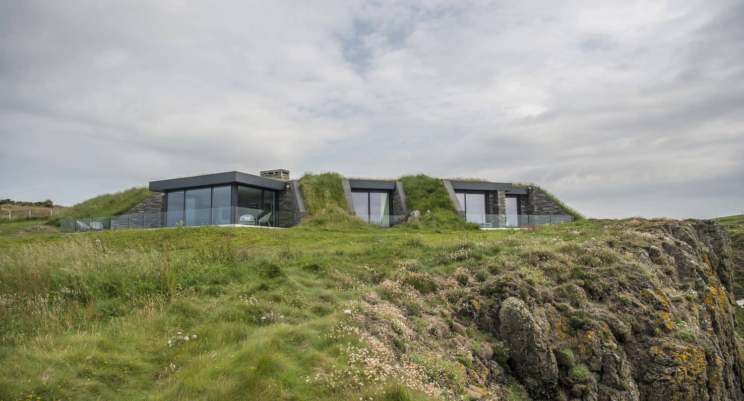 A modern house with large glass windows and sleek bifold doors is built into a grassy hillside, blending with the landscape. The building overlooks rocky cliffs and is surrounded by lush green grass under a cloudy sky.
