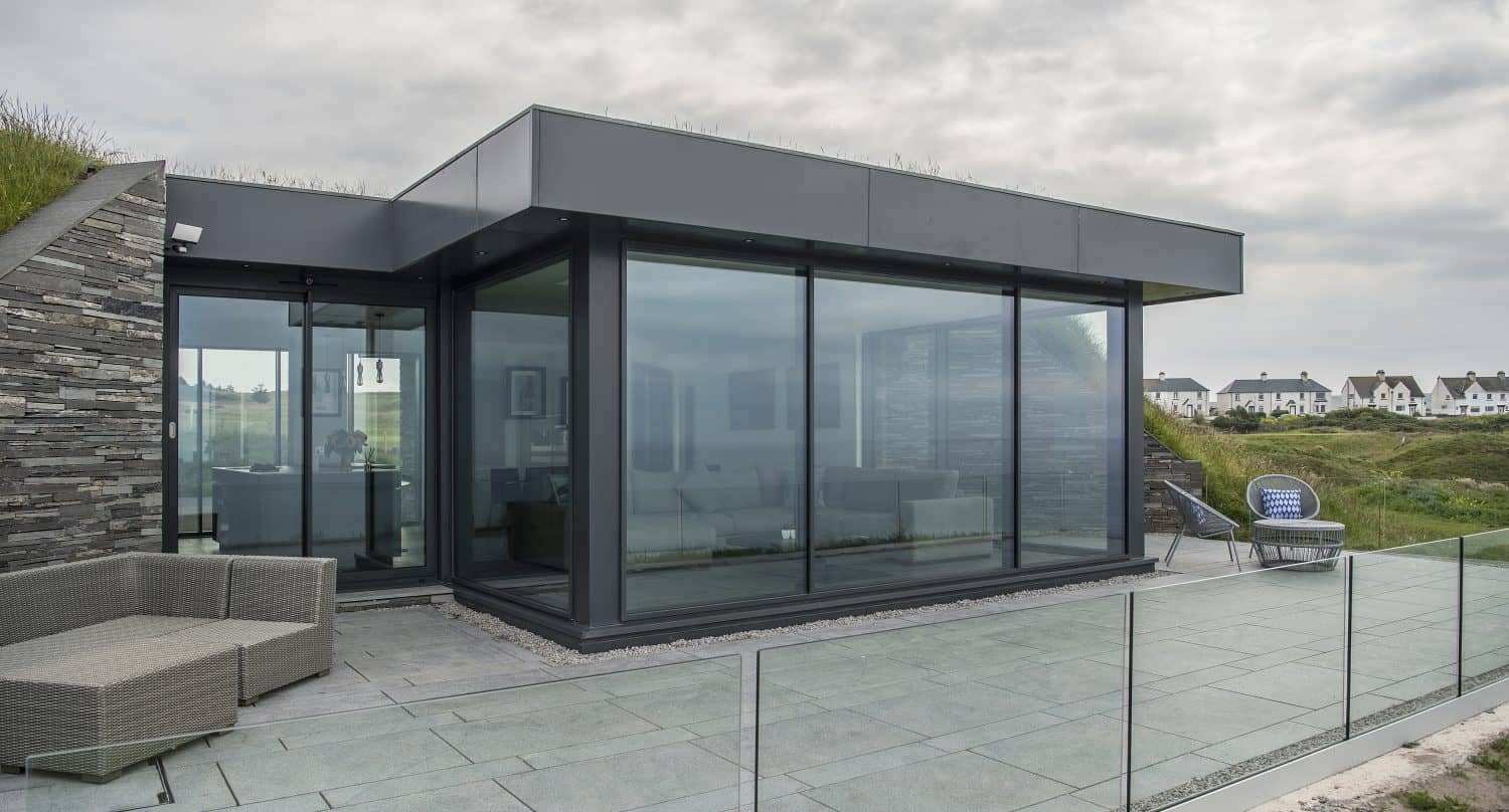 Modern glass-walled house featuring bespoke glass solutions, with a patio, outdoor seating, and glass railing, set against a grassy landscape under a cloudy sky. Houses can be seen in the distance.