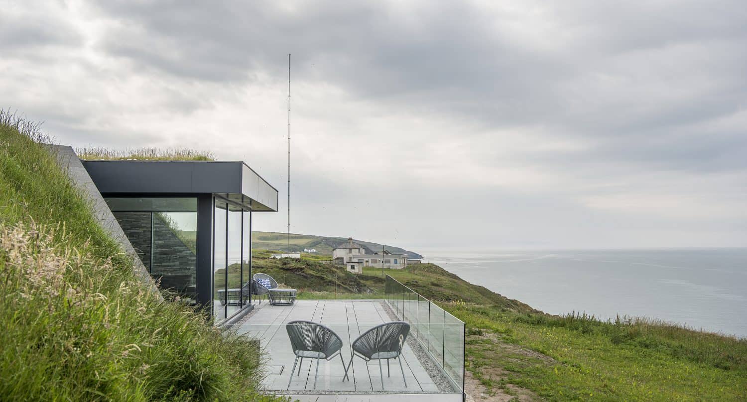 A modern glass-walled building with sleek sliding doors and a grassy roof overlooks the ocean from a hilltop. Two black chairs sit on a patio with a glass railing, surrounded by green grass and distant coastal houses.
