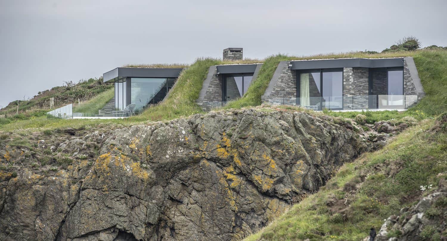 Modern houses with large glass windows and sliding doors are built into a grassy hillside, overlooking a rugged cliff edge with patches of moss and vegetation, under an overcast sky.