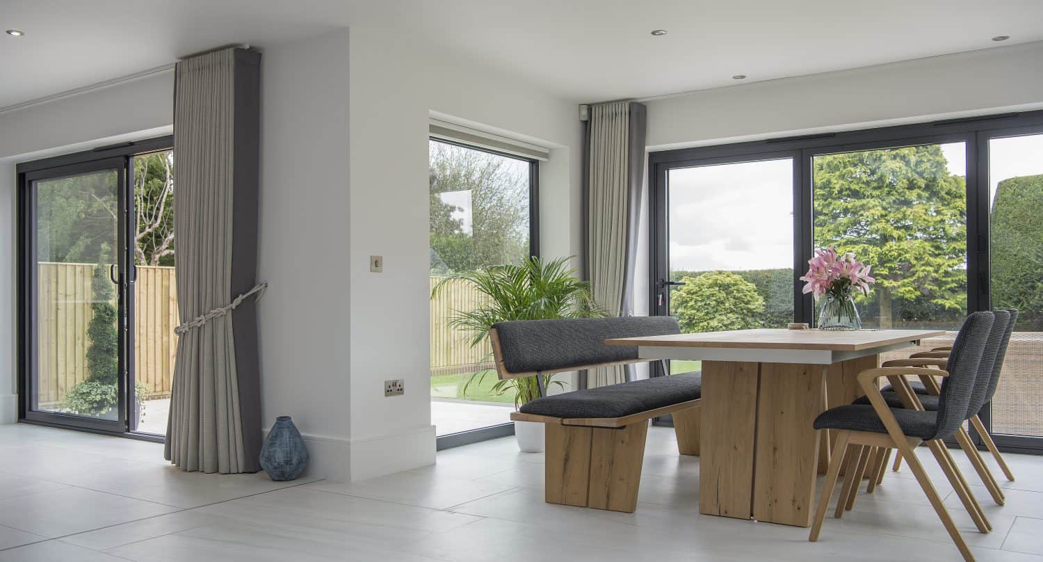 Modern dining area with a wooden table, gray benches, and pink flowers in a vase. Large windows and bespoke glass solutions like sliding doors offer views of a green garden, filling the space with natural light and minimalist charm.