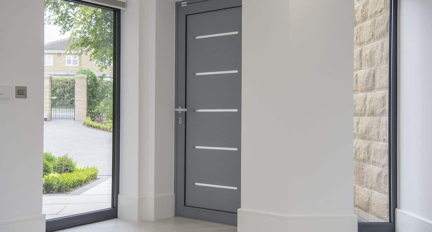 Modern gray front door with horizontal glass panels, surrounded by large floor-to-ceiling windows, showcases bespoke glass solutions that allow natural light into a bright, white entryway with views of a garden and driveway outside.