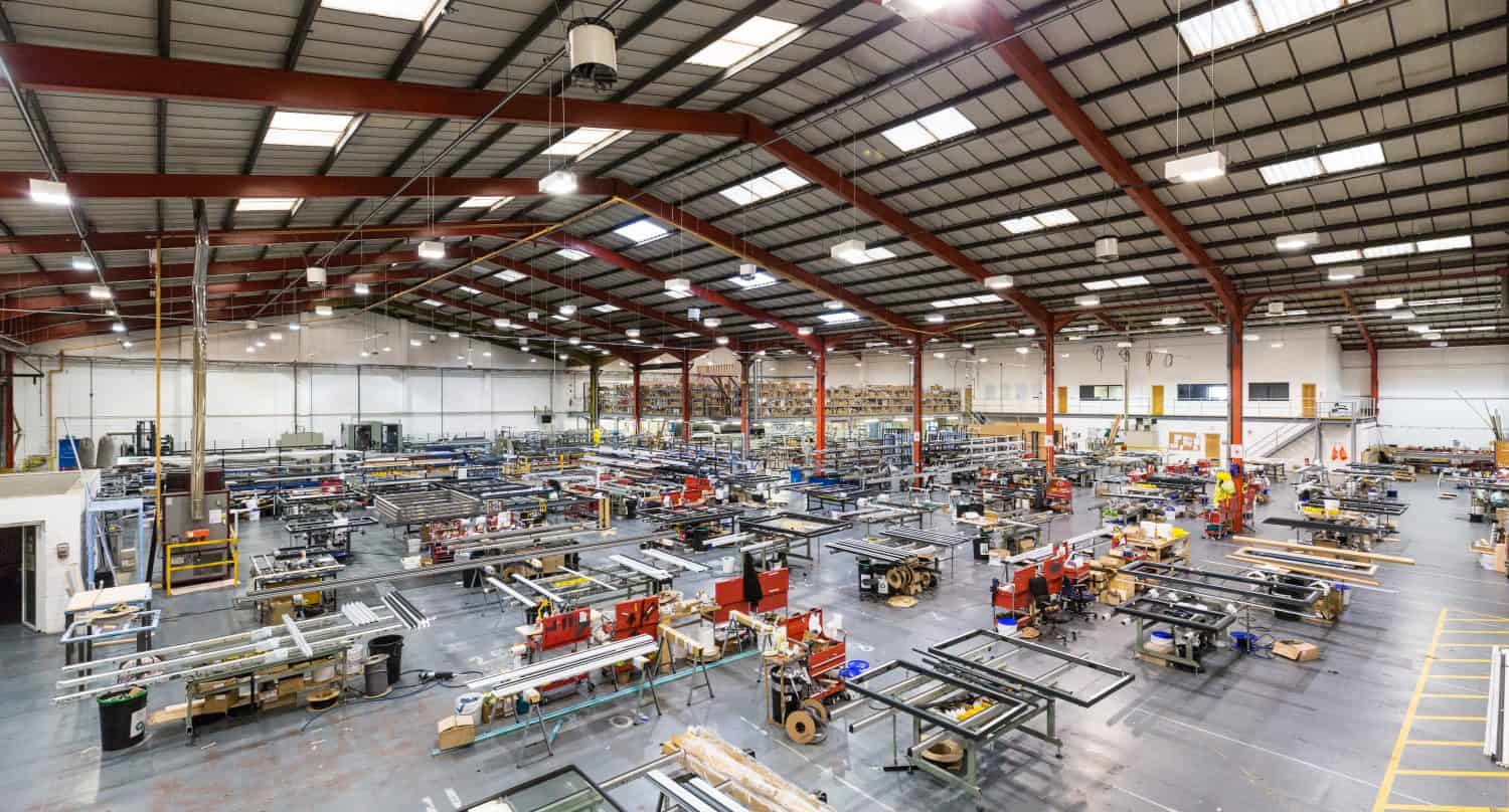 Wide view of a large, well-lit manufacturing factory with high ceilings, exposed beams, and multiple workstations filled with industrial equipment and metal frames, featuring glass roofing that floods the organized rows with natural light.