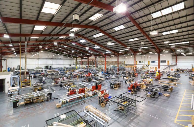 Wide view of a large, well-lit manufacturing factory with high ceilings, exposed beams, and multiple workstations filled with industrial equipment and metal frames, featuring glass roofing that floods the organized rows with natural light.