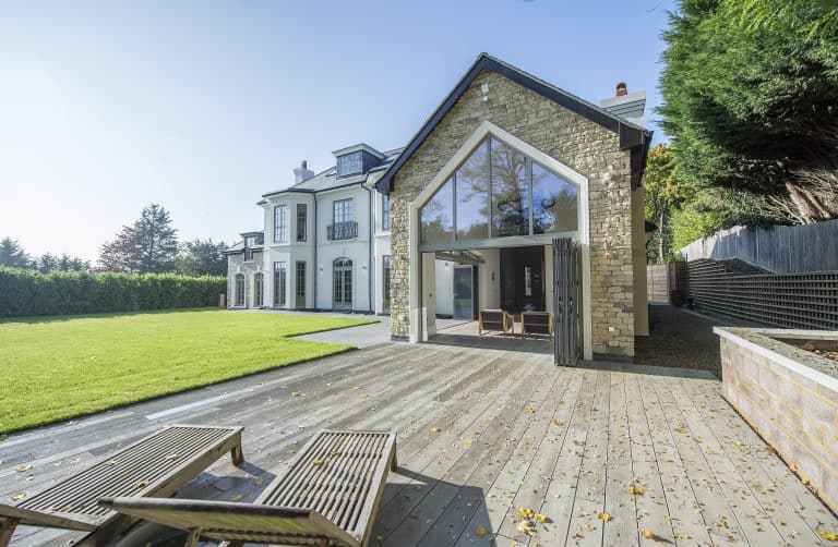 Large modern house with tall windows, stone facade, and stylish bifold doors, adjacent to a spacious wooden deck with lounge chairs. The deck opens to a green lawn bordered by trees and a wooden fence under a clear blue sky.