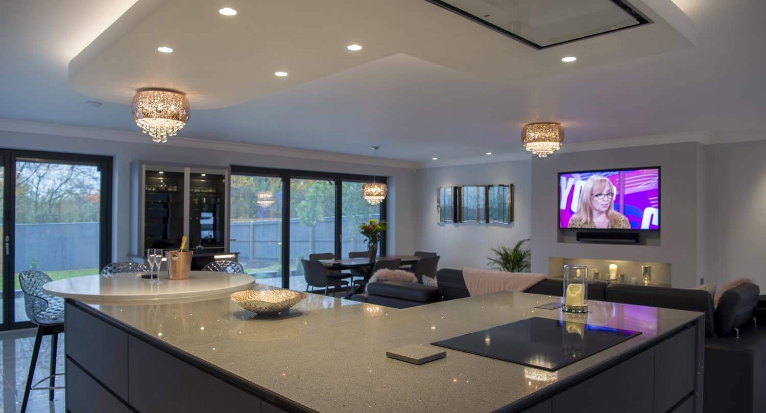 Modern open-plan kitchen and living area with large island, bar stools, sofas, dining table, and a wall-mounted TV showing a woman. Bespoke glass solutions feature in the glass doors leading to the balcony; chandeliers hang from the ceiling.