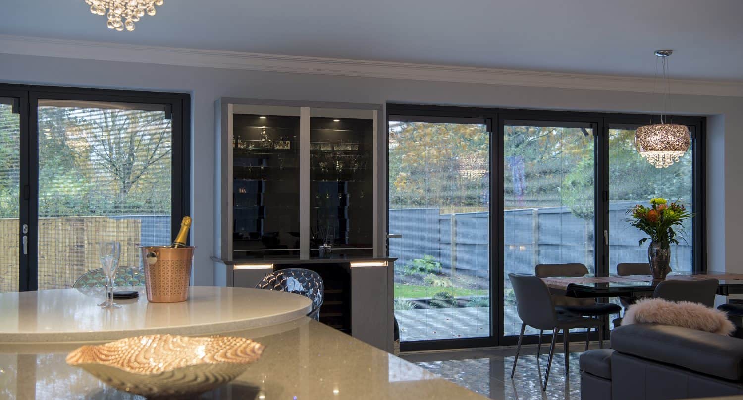 Modern kitchen and dining area with large bifold doors overlooking a garden. A round island counter with a decorative bowl and champagne bucket sits beneath two chandeliers, creating a warm ambiance.