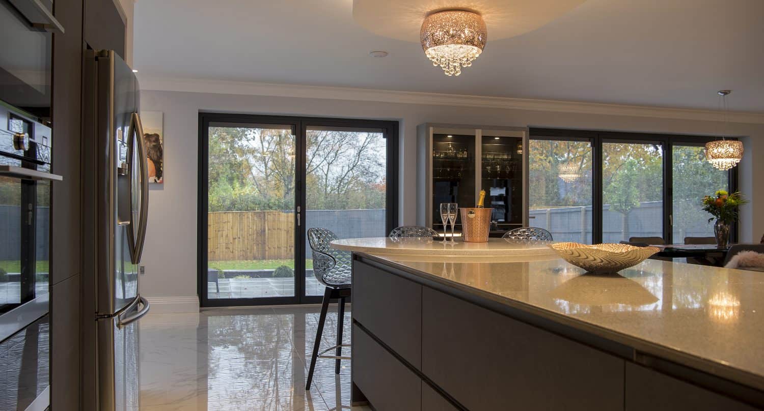 Modern kitchen with a large island, barstools, elegant pendant lights, and floor-to-ceiling windows offering a view of a fenced backyard with trees. Sliding doors enhance the space, while champagne glasses and a decorative bowl sit on the island.