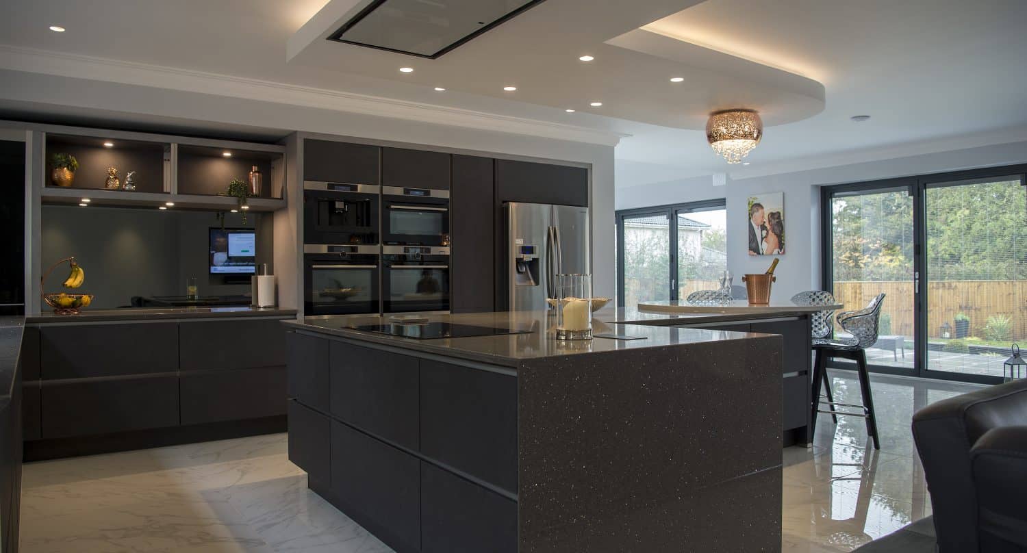 Modern kitchen with dark cabinets, a large central island, built-in appliances, and a stainless steel fridge. Floor-to-ceiling bifold doors let in natural light, and there is a small dining area near the windows.
