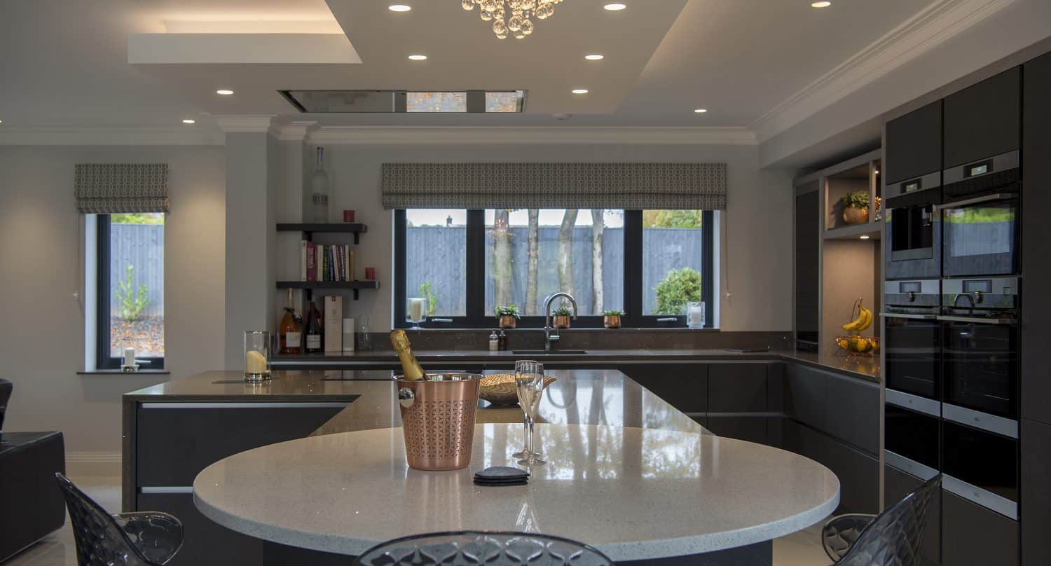Modern kitchen with dark cabinets, a large round island, built-in ovens, and a chandelier overhead. Bespoke glass solutions in the windows let in natural light while a champagne bottle chills on the island.