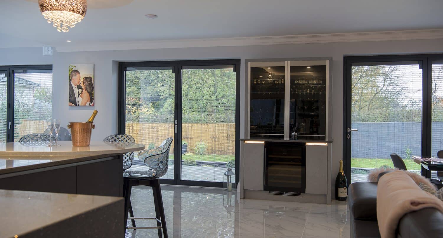 Modern kitchen and living area with large sliding doors overlooking a garden, marble floors, a kitchen island with a champagne bucket, seating area, photo on the wall, and visible wine fridge.