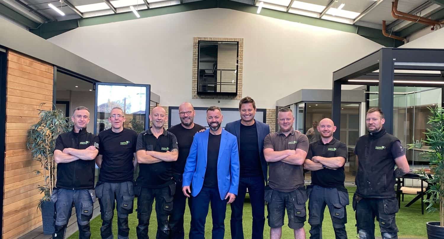 Nine men stand side by side in a modern indoor space with artificial grass, framed by sleek bifold doors. Eight wear black work clothes, while the man in the center sports a bright blue blazer. They all smile or pose for the camera.