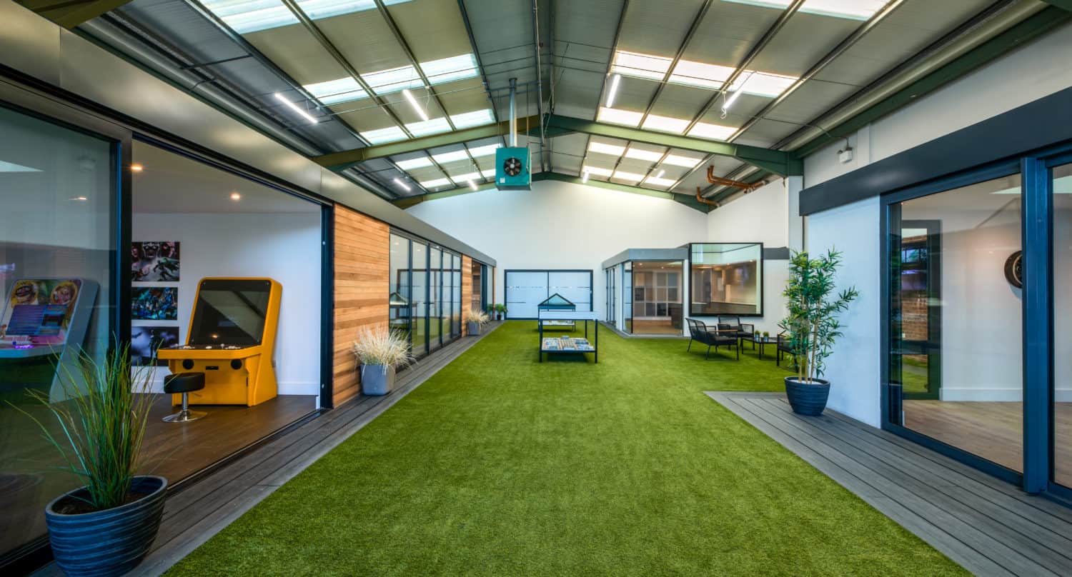 A modern indoor space with a green artificial grass runway down the center, surrounded by bespoke glass solutions, potted plants, tables, chairs, and an arcade machine. The high ceiling features sleek skylights for abundant natural light.