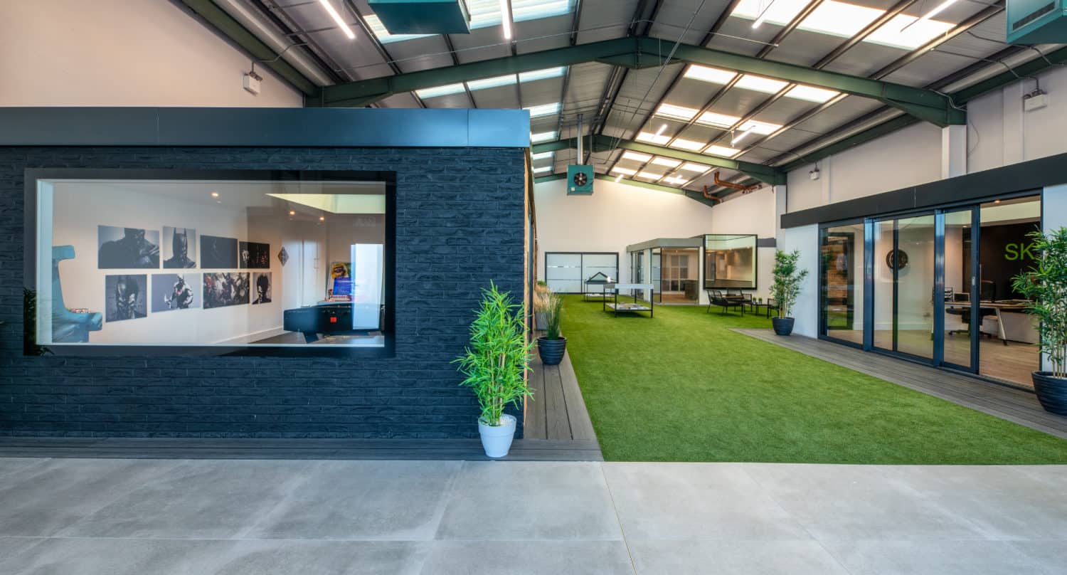 Modern indoor space with high ceilings, artificial grass, potted plants, and large windows. Glass roofing enhances the bright, spacious central area, bordered by office rooms and finished with industrial lighting and decor.