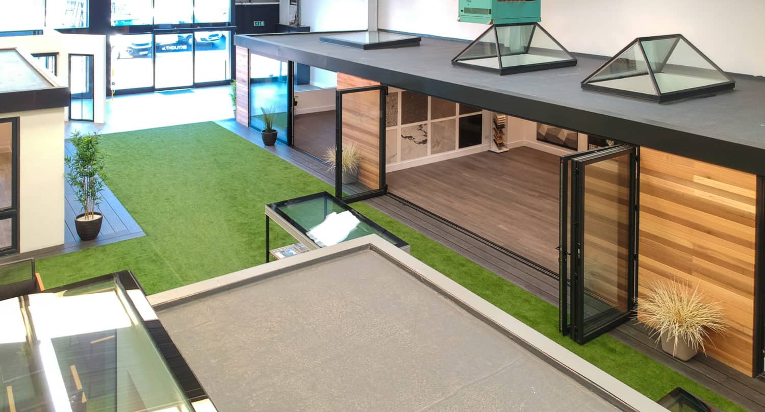 Spacious modern indoor showroom with artificial grass flooring, large windows, wooden structures, bifold doors, potted plants, and natural light flooding in from bespoke glass solutions at the wide entrance.