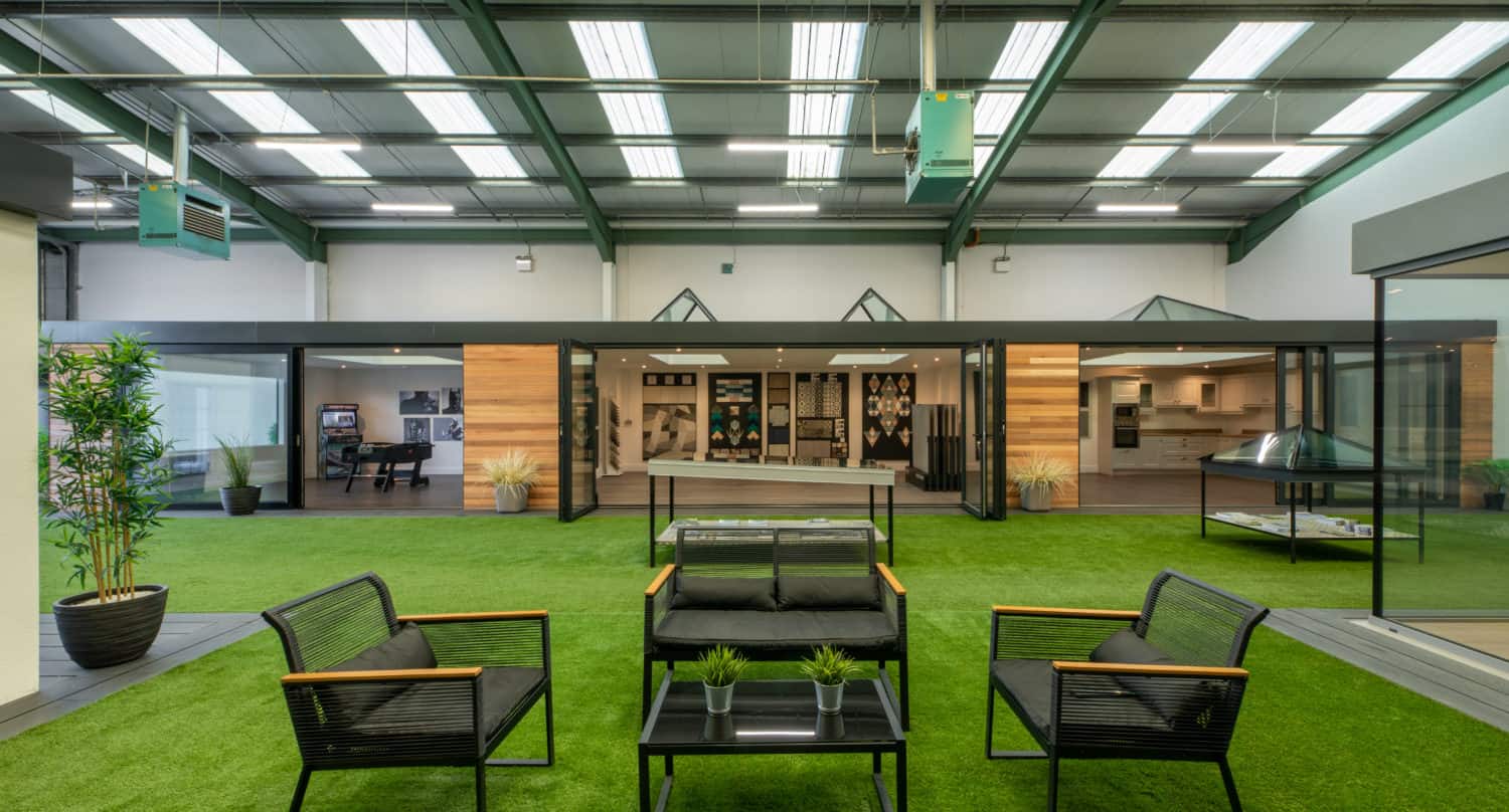 A modern indoor space with artificial grass flooring, black patio furniture with wooden armrests, potted plants, and a glass-walled building featuring bespoke glass solutions under a high, brightly lit ceiling.