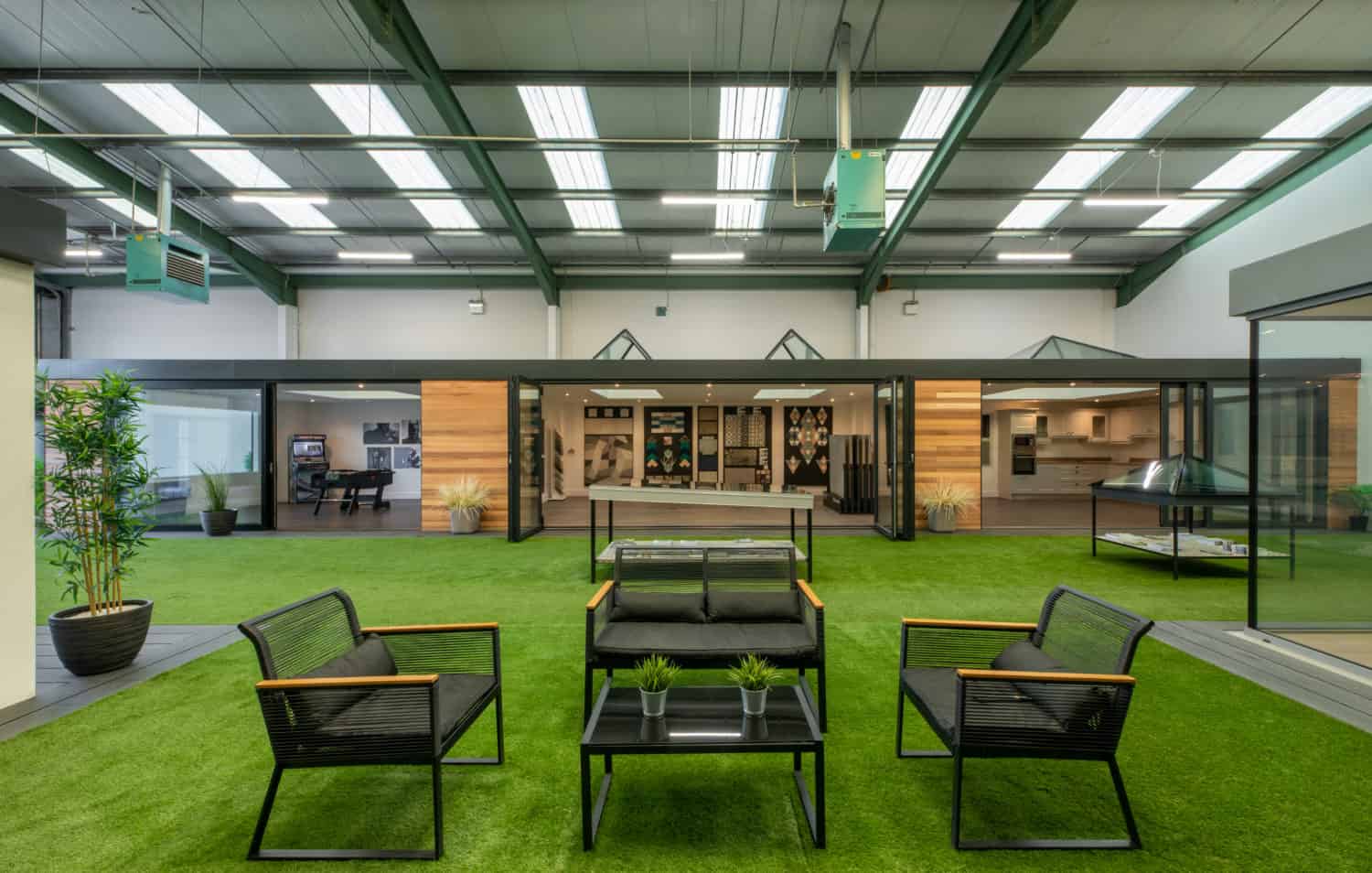 A modern indoor space with artificial grass flooring, black patio furniture with wooden armrests, potted plants, and a glass-walled building featuring bespoke glass solutions under a high, brightly lit ceiling.