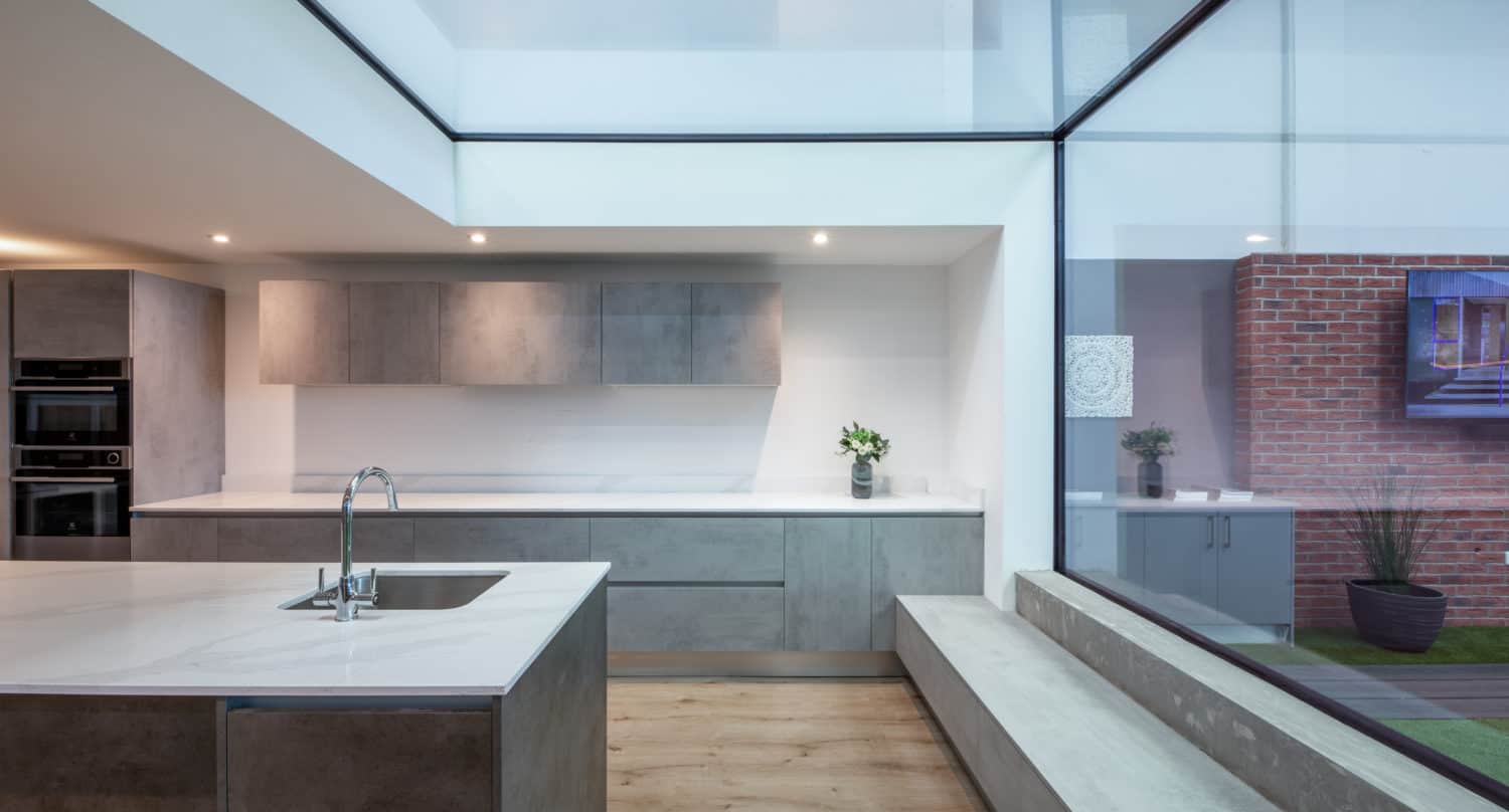 Modern kitchen with minimalist gray cabinets, a white island and sink, light wood flooring, large windows, and a built-in bench by the glass wall with sliding doors opening to a brick patio with plants.