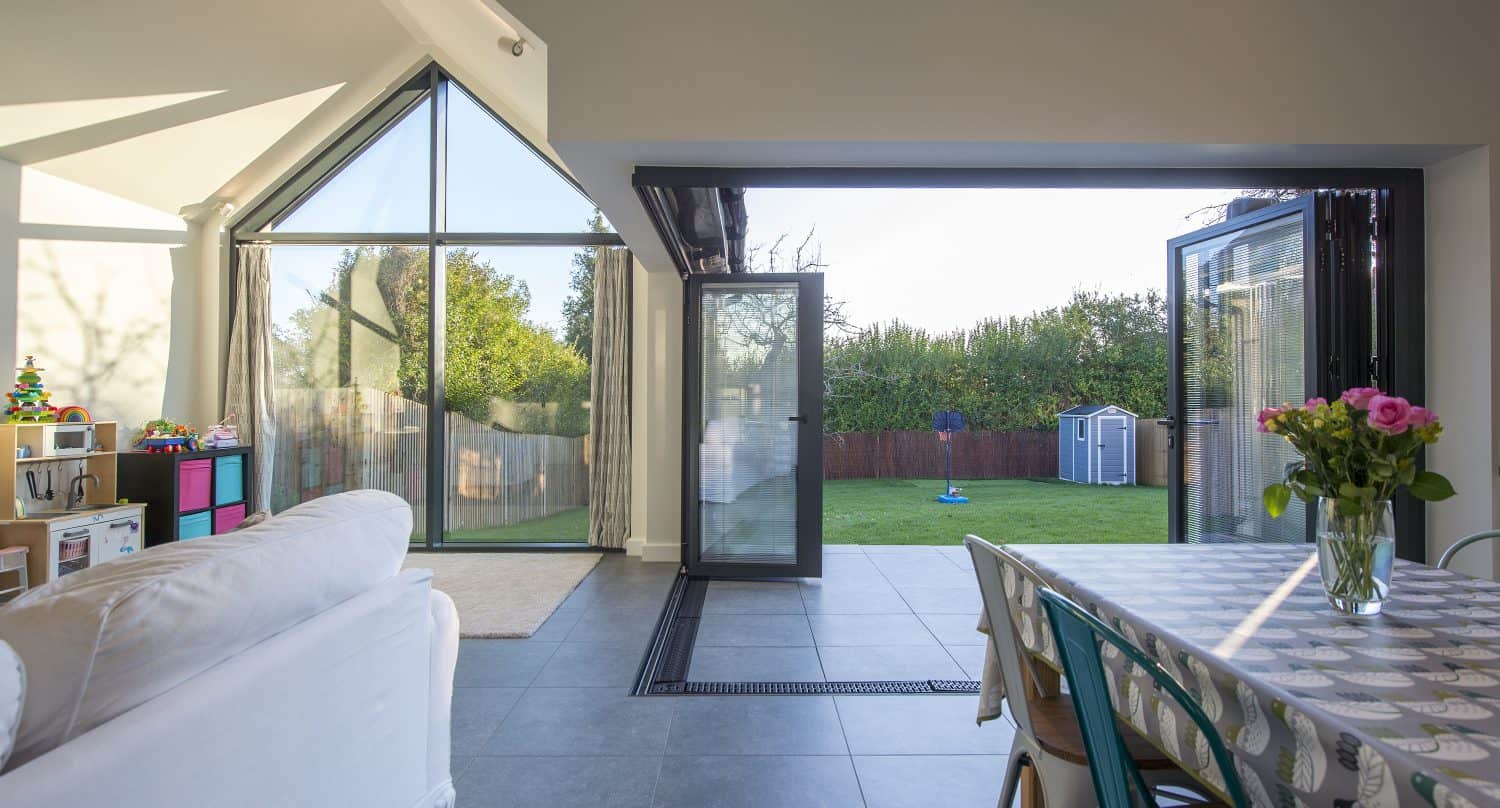 Bright, modern living space with large bifold doors open to a green backyard; a sofa, children’s toys, and a dining table with flowers are visible inside. Natural light fills the room.