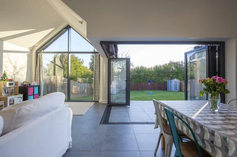 Bright, modern living space with large bifold doors open to a green backyard; a sofa, children’s toys, and a dining table with flowers are visible inside. Natural light fills the room.