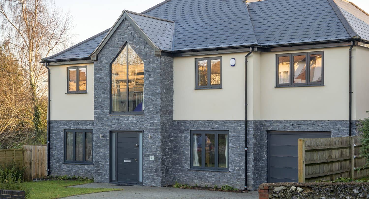 Modern two-story house with cream walls, gray stone accents, large windows, and a triangular central window above the main door. Sliding doors open to the driveway with a small grassy area and wooden fencing. Slate roof tops the stylish design.