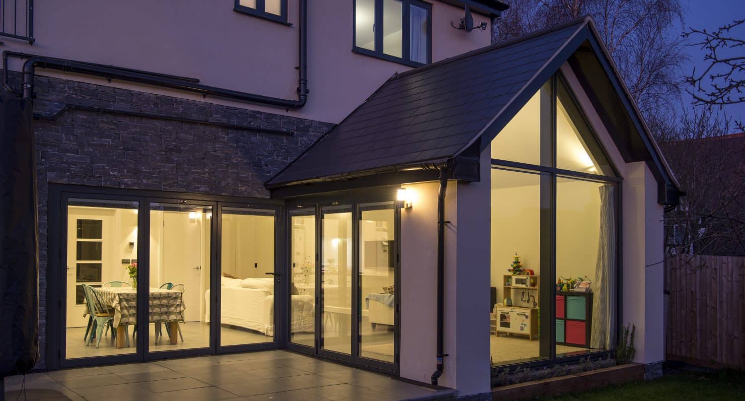 Modern house at dusk with bespoke glass solutions, featuring large bifold doors and windows. The warmly lit interior reveals a dining area, living space, childrens playroom, plus patio tiles and a garden outside.