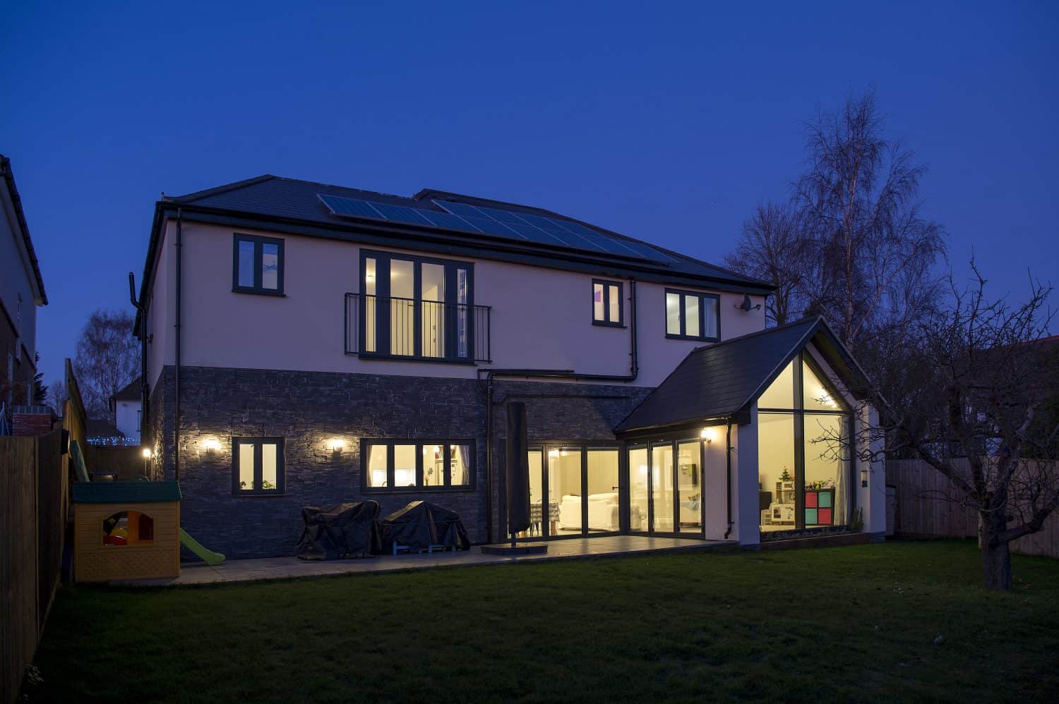 A modern two-story house with large windows and bifold doors is illuminated at dusk. Solar panels are visible on the roof, and the backyard includes a small playhouse and a lawn bordered by a fence and trees.