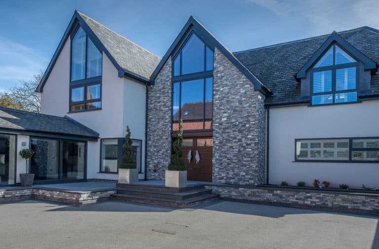 Modern two-story house with large glass windows, brick and white exterior, stylish bifold doors at the entrance, sculpted topiary plants by the doorway, and a paved driveway under a blue sky.