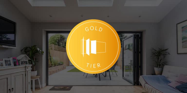 A bright, modern living room with bifold doors opening to a patio and garden, overlaid with a gold circular badge reading Gold Tier and a stylized folding door icon in the center.
