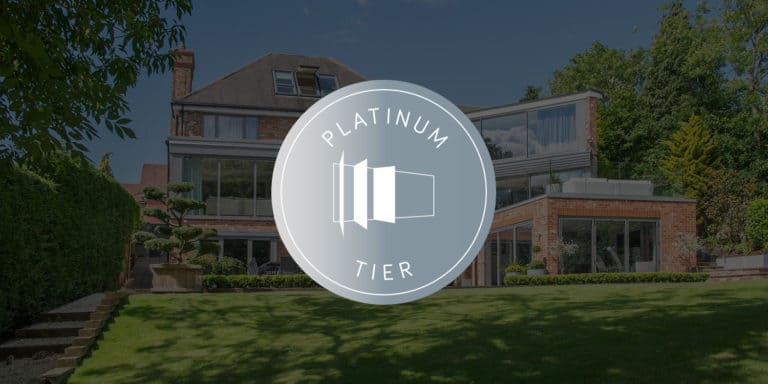 A large, modern brick house with bifold doors and a spacious lawn, partially covered by a circular Platinum Tier badge featuring a stylized icon in the center.