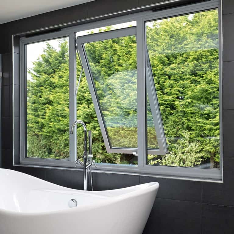 A modern bathroom features a white freestanding bathtub in front of large, gray-framed sliding doors, one of which is open to reveal lush green trees outside.