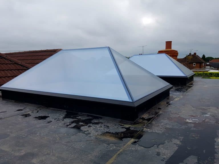 Two modern pyramid-shaped skylights, crafted as part of bespoke glass solutions, sit on a flat rooftop. With frosted glass panels and dark bases, they stand out against cloudy skies and nearby houses in the background.