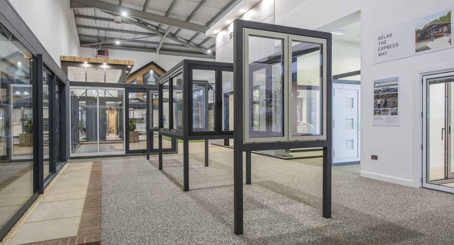 A modern showroom displays various large glass windows, sliding doors, and black-framed front doors on stands in a bright, spacious, industrial-style interior with high ceilings.