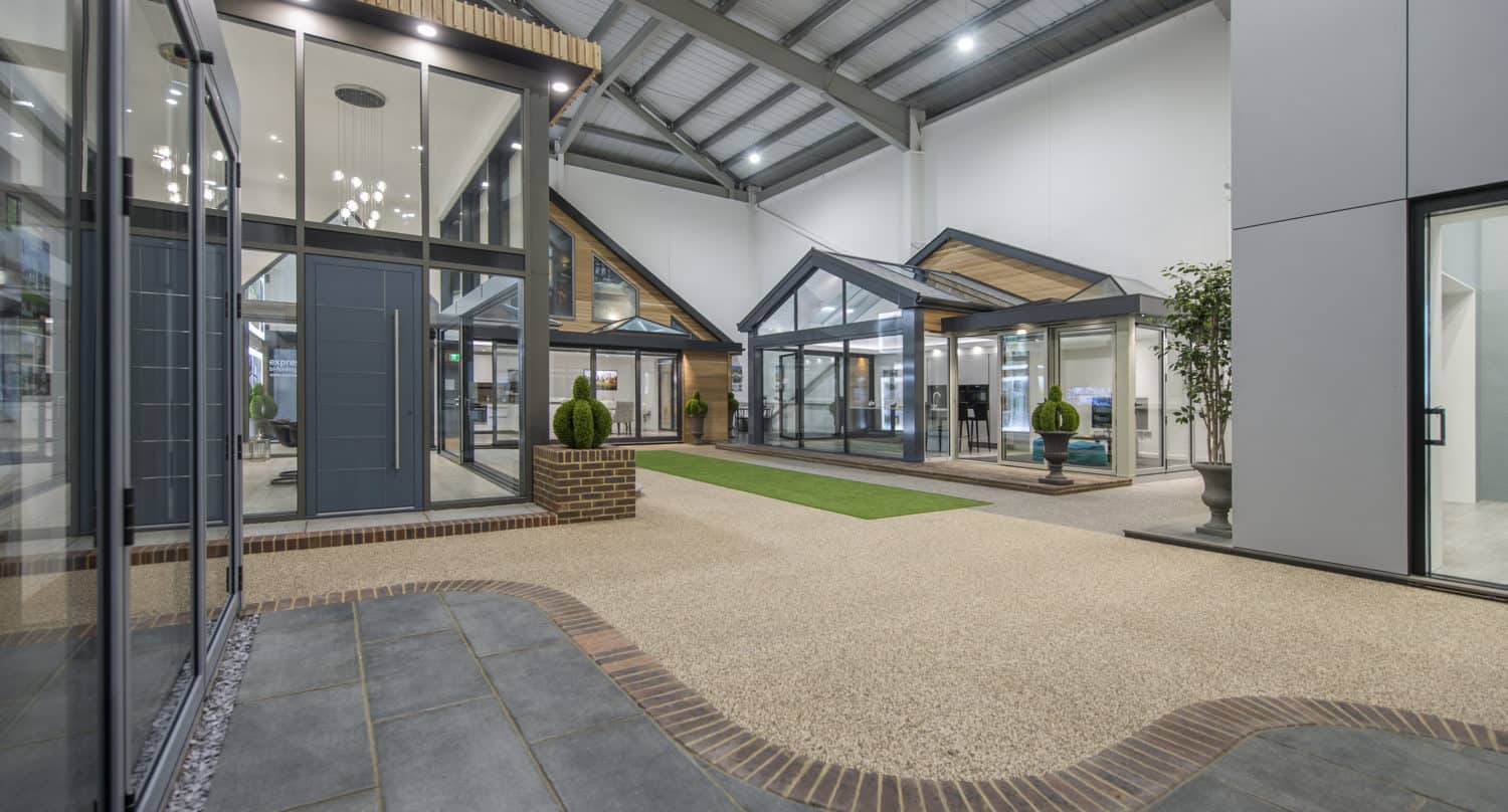 A modern indoor showroom displays various glass doors, front doors, windows, and conservatory structures under a high metal ceiling, with potted plants, tiled paths, and artificial grass.