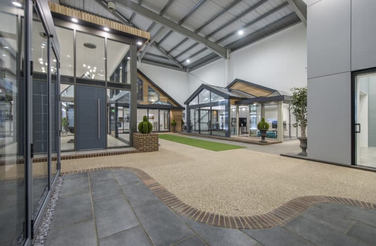Indoor showroom featuring modern glass and metal house facades with large windows, sliding doors, pitched roofs, potted plants, and a clean, open layout with tiled and gravel pathways under a high ceiling.