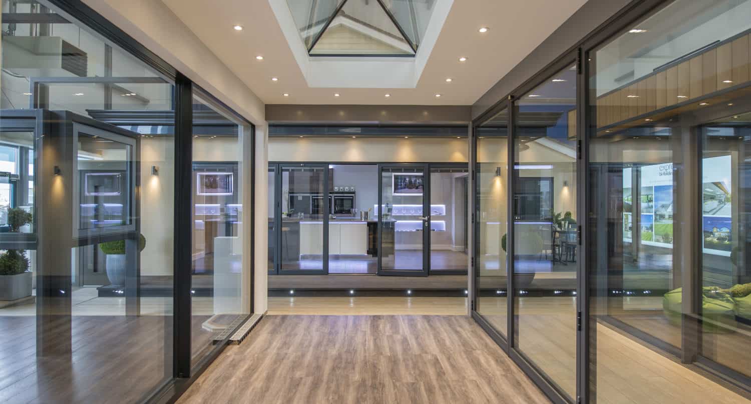 A modern indoor hallway with glass walls, wooden flooring, recessed ceiling lights, a skylight, and glass roofing, leading to a spacious living area and kitchen in the background.