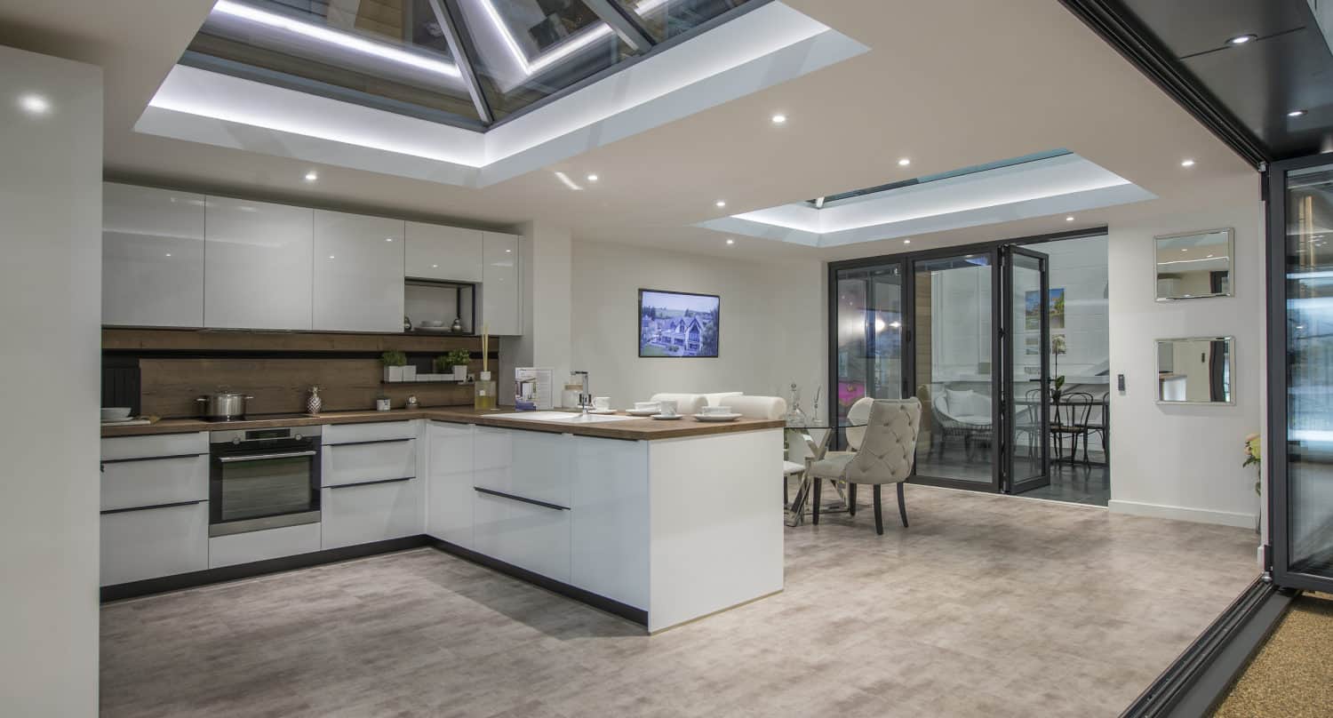 Modern kitchen with white cabinets, a large island, and built-in appliances. The space features glass roofing and an open dining area with a table and chairs, connected to a patio through sleek sliding doors.