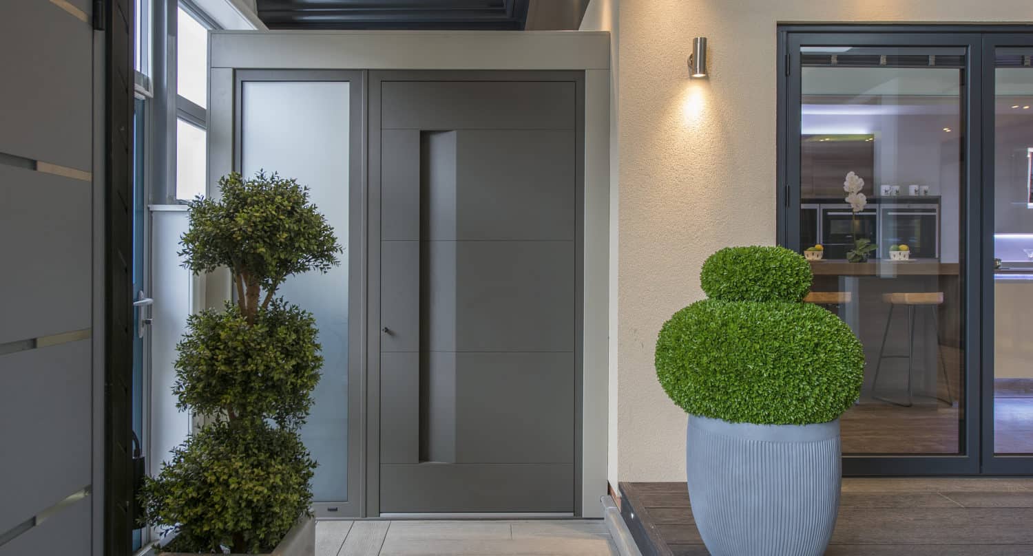 Modern house entrance with a sleek gray front door and two large potted topiary plants. Clean lines, a light-colored wall, and bespoke glass solutions in the large sliding doors reveal a contemporary interior.