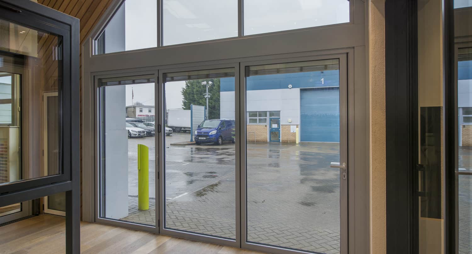 Large glass sliding doors open to a wet, paved area outside with parked cars, a blue roller door, and industrial buildings visible through the windows on a rainy day beneath partial glass roofing.
