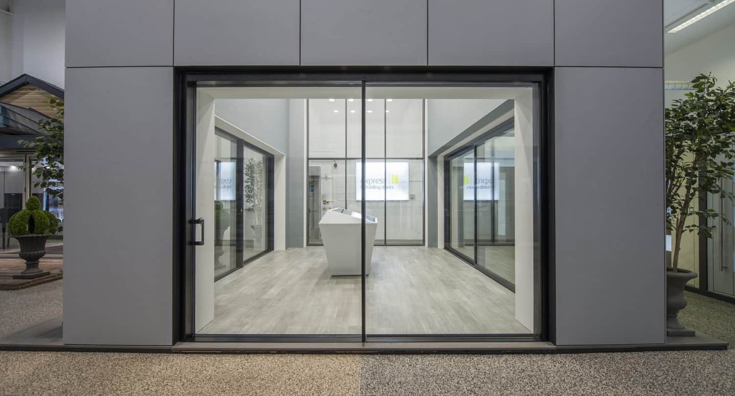 A modern building entrance with large glass sliding doors, gray panels, and light wood flooring inside. Two small indoor trees are visible on either side of the front doors.